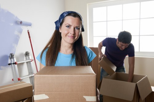 A woman with long brown hair, wearing a blue shirt and a blue headscarf, stands in a bright room with white walls and large window panes, holding a cardboard box on top of a stack of similar boxes. To her right, a man with dark curly hair, dressed in a navy-blue T-shirt, is bending down while placing or adjusting another cardboard box, with additional boxes scattered around on the floor. The room appears to be a residential interior, possibly an entrance hallway or living area, with natural light illuminating the scene. Visible house removal supplies such as packing materials, cardboard boxes, and minor items suggest an ongoing move. The setting indicates a professional house removals service, typical of Battersea Man with Van, with the focus on packing and preparing household belongings for relocation within the Battersea area. The furniture includes cardboard boxes in various sizes, possibly filled with personal items, all positioned centrally in the room, with some boxes partially opened. The overall atmosphere is organized and professional, emphasizing careful packing and handling during a residential move.
