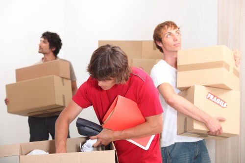 Three men involved in a house removal process inside a residential property. The man in the foreground, wearing a red t-shirt, is bent over, lifting a cardboard box while holding a red folder and a black device. Behind him, a man in a white t-shirt is carrying a medium-sized cardboard box labeled 'FRAGILE' towards the right side of the image. To the left, another man with dark, curly hair, dressed in a grey t-shirt, is in the background holding a larger cardboard box, looking towards the left. The room has light-colored walls, with a window partially visible on the right, covered by a white curtain. The scene depicts typical house removals with moving boxes of various sizes, showing the physical effort involved in packing and transporting household items, consistent with professional removal services in Battersea or nearby Wandsworth areas. The environment is well-lit, with a clear focus on the transferring of household contents, highlighting elements relevant to house removals and logistical planning by Battersea Man with Van.